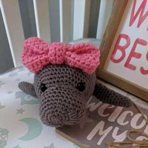 Puede incluir: Una manat&iacute; de crochet gris con un lazo rosa en la cabeza. La manat&iacute; est&aacute; acostada sobre una manta blanca y verde con estrellas y lunas.
