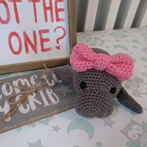 Puede incluir: Un juguete de peluche de manat&iacute; de crochet gris con un lazo rosa. El juguete est&aacute; acostado sobre una tela estampada de color blanco y verde con el texto "Come to my crib" en un letrero de madera detr&aacute;s de &eacute;l.