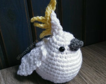 May include: A crocheted white cockatoo with a yellow crest. The cockatoo has black eyes and a gray beak.