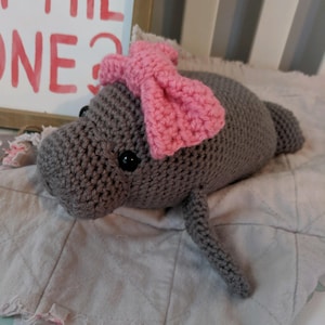 Puede incluir: Un manat&iacute; de crochet gris con un lazo de crochet rosa en la cabeza. El manat&iacute; est&aacute; acostado sobre una manta blanca y gris.