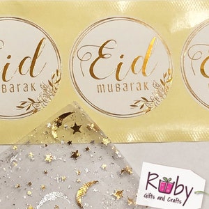 May include: Gold foil stickers with the text "Eid Mubarak" and "Eid Barak" on a yellow background. The stickers are next to a white sheer fabric with gold stars and crescent moons.