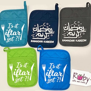 May include: Five different pot holders with various designs. Two pot holders are blue with the text "Is it iftar yet?!" and a fork and knife. One pot holder is green with the same text and a fork and knife. One pot holder is dark blue with Arabic text and the words "Ramadan Kareem". One pot holder is gray with the same Arabic text and the words "Ramadan Kareem".