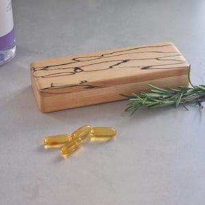 May include: A rectangular wooden block with natural light brown tones and dark, irregular spalted grain patterns. Beside the block lies a small sprig of green rosemary and four translucent, golden-yellow oval gel capsules.