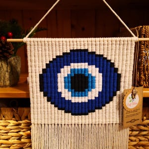 May include: A macrame wall hanging featuring a blue and white evil eye design. The eye is centered on a cream-colored background, with a fringe at the bottom. The wall hanging is suspended from a wooden dowel.
