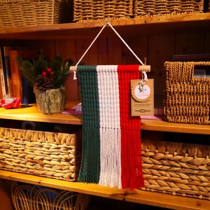 以下が含まれることがあります： 緑、白、赤のカラーブロックデザインのマクラメ壁掛け。木製のダボから吊り下げられています。壁掛けは、編まれたバスケットや装飾品のある木製の棚に飾られています。
