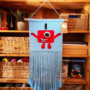 May include: A light blue macrame wall hanging featuring a red cartoon character with a black top hat. The wall hanging has a fringe at the bottom and is suspended from a wooden dowel with a white string.