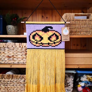 May include: A macrame wall hanging featuring a pixelated jack-o'-lantern design in orange, black, and yellow on a light purple background. The piece is suspended from a wooden dowel by a black cord and includes a long, yellow fringe at the bottom.