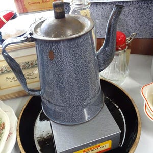 May include: A vintage gray speckled enamel coffee pot with a matching lid, a curved handle, and a long spout. The pot sits atop a gray metal Kodaslide Compartment File box, which is placed inside a large, round, black ceramic bowl with a gold rim.