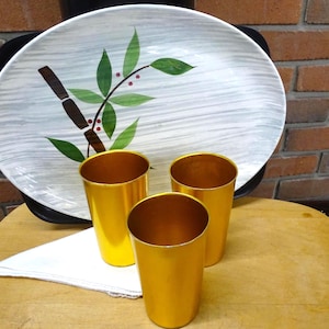 May include: An oval serving platter with a white background, grey brushstroke textures, and a green leaf and brown bamboo-style stem design. Three cylindrical gold-colored metal tumblers are arranged in front of the platter on a white cloth.