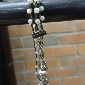 May include: A multi-strand bracelet featuring white beads and brown cord. The bracelet has a silver clasp and chain with heart and flower charms. The background is a brick wall.