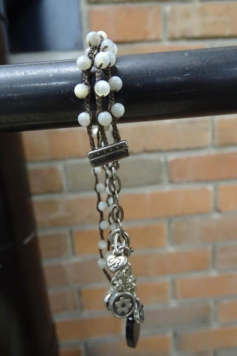 May include: A multi-strand bracelet featuring white beads and brown cord. The bracelet has a silver clasp and chain with heart and flower charms. The background is a brick wall.