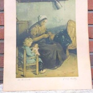 Puede incluir: Una impresión enmarcada de una escena doméstica, que presenta a una mujer sentada y trabajando, con un niño sentado cerca. La impresión tiene una paleta de colores apagados con el texto "At the Cradle" y una firma en la parte inferior.