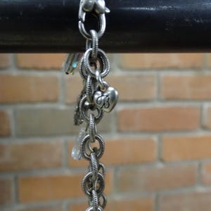 May include: Silver-toned bracelet with multiple charms, including a heart-shaped clasp and a heart charm with the letter "B". The bracelet is made of linked rings and hangs against a brick wall.