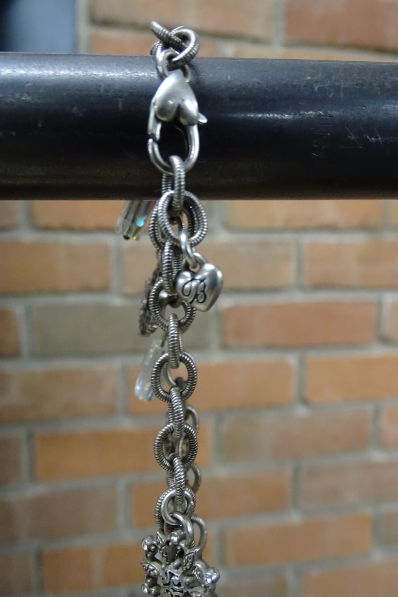 May include: Silver-toned bracelet with multiple charms, including a heart-shaped clasp and a heart charm with the letter "B". The bracelet is made of linked rings and hangs against a brick wall.