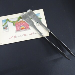 Op de afbeelding: Twee zilveren botermesjes met sierlijke handvatten. De mesjes liggen op een vintage kerstkaart met de tekst "A Hearty New Year".