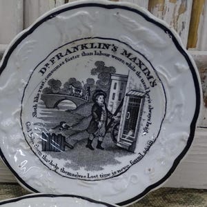 May include: A white plate with a decorative, scalloped edge and a black rim. The plate has a black and white illustration of a boy with a cart, a bridge, and text that reads "Dr. Franklin's Maxims".