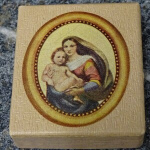 May include: A small wooden box with a gold oval frame containing a painting of the Virgin Mary holding a baby Jesus. The painting is on a beige background.