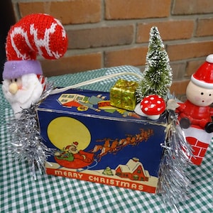 Puede incluir: Una decoración navideña con una figura de Papá Noel, un pequeño árbol de Navidad y un hongo rojo y blanco. Las figuras se colocan encima de una caja de estilo vintage con fondo azul y las palabras "Feliz Navidad" impresas en la parte delantera.