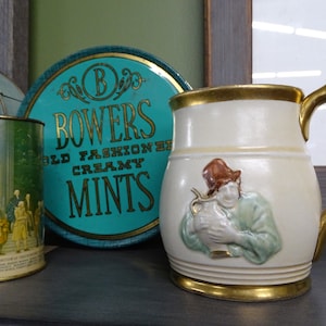 Può includere: Due scatole rotonde di menta color turchese con lettere dorate che recitano "Bowers Old Fashioned Creamy Mints" e una tazza in ceramica bianca con un bordo e un manico dorati. La tazza ha un'immagine in rilievo di un uomo con una tunica verde che tiene un'anfora.