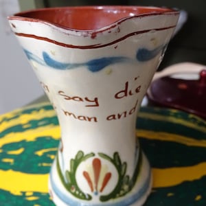 Puede incluir: Un jarrón de cerámica blanca con un borde rojo y diseños florales azules y verdes. El jarrón tiene las palabras "Say die man and" escritas en él.