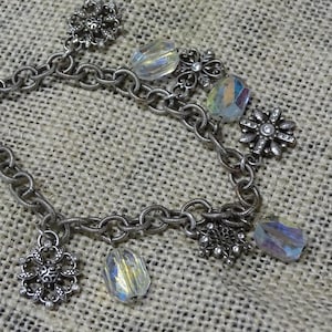 May include: Silver-toned bracelet with a chain link design. The bracelet features several decorative elements, including clear, iridescent crystal beads and ornate, flower-shaped charms. The bracelet is displayed on a textured, neutral-colored surface.