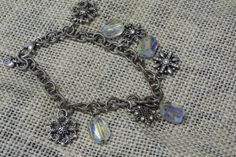 May include: Silver-toned bracelet with a chain link design. The bracelet features several decorative elements, including clear, iridescent crystal beads and ornate, flower-shaped charms. The bracelet is displayed on a textured, neutral-colored surface.
