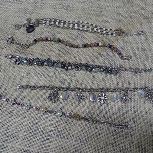 May include: Five silver-toned bracelets with various designs are displayed on a textured surface. One bracelet features white beads, another has colorful beads, and a third has blue and clear stones. The remaining two bracelets have charms, including snowflakes and iridescent teardrops.