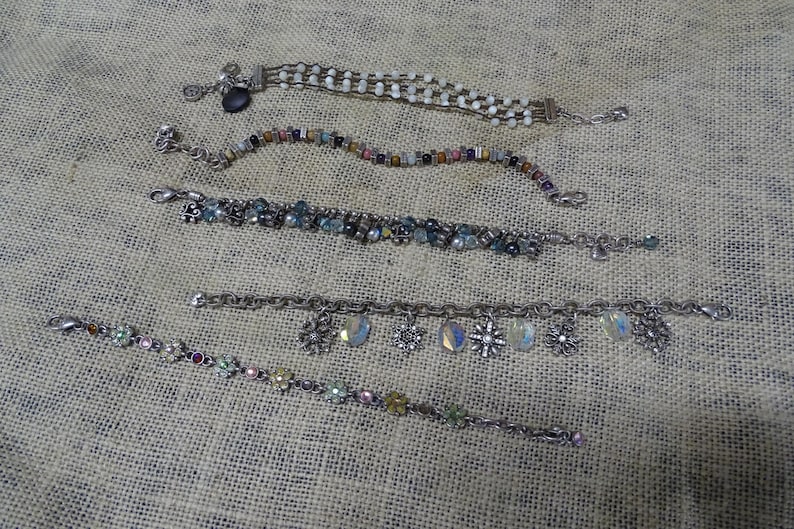 May include: Five silver-toned bracelets with various designs are displayed on a textured surface. One bracelet features white beads, another has colorful beads, and a third has blue and clear stones. The remaining two bracelets have charms, including snowflakes and iridescent teardrops.