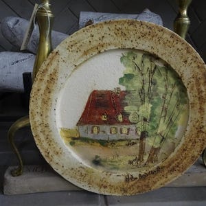 May include: A decorative ceramic plate featuring a hand-painted scene of a house with a red roof and green trees. The plate has a speckled, textured rim and a cream-colored center. The artwork is signed in the lower right corner.