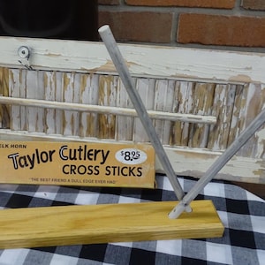May include: A wooden knife sharpening tool with two angled cross sticks, mounted on a wooden base. The product is from Taylor Cutlery, with the text "CROSS STICKS" and "THE BEST FRIEND A DULL EDGE EVER HAD" on the packaging.