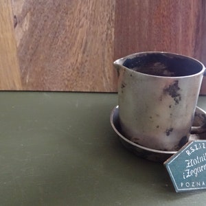 Puede incluir: Una pequeña cremera plateada deslustrada con asa y platillo adjunto. Una etiqueta con texto en polaco cuelga del asa. La cremera está sobre una superficie verde con un fondo de madera.