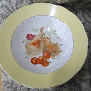 May include: A white ceramic bowl with a yellow rim and a painted design of pears and cherries. The bowl is on a gray and white fabric.