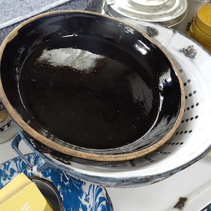 May include: An oval, black ceramic baking dish with a brown rim sits inside a white and blue enamelware colander. A glass jar of coffee and other vintage kitchenware are in the background.