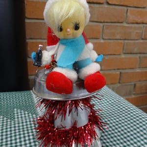 May include: A vintage Christmas tree decoration featuring a small doll wearing a white hat, blue scarf, and red boots. The doll is sitting on a silver metal base with red tinsel around it.