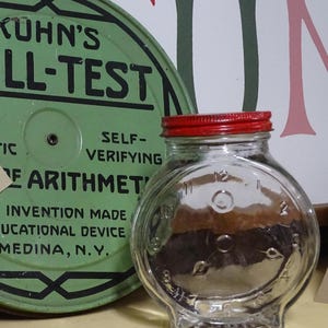 Puede incluir: Un tarro de vidrio transparente con tapa roja, con forma de esfera de reloj, con los números del 1 al 12. En el fondo, un letrero metálico verde con texto negro que dice "KUHN'S LL-TEST". El letrero también dice "SELF-VERIFYING" y "EARITHMET".