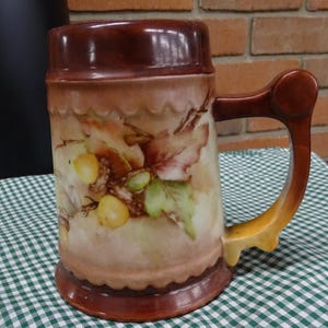 Pode incluir: Uma caneca de cerveja de cerâmica marrom e branca com uma alça de madeira. A caneca é decorada com um desenho pintado de folhas e bagas.