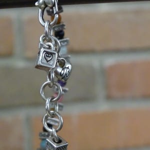 May include: Silver-toned charm bracelet with heart and square charms, and colorful beads. The bracelet features a variety of charms, including hearts and squares, and is strung with a mix of colorful beads. The bracelet is displayed against a brick background.