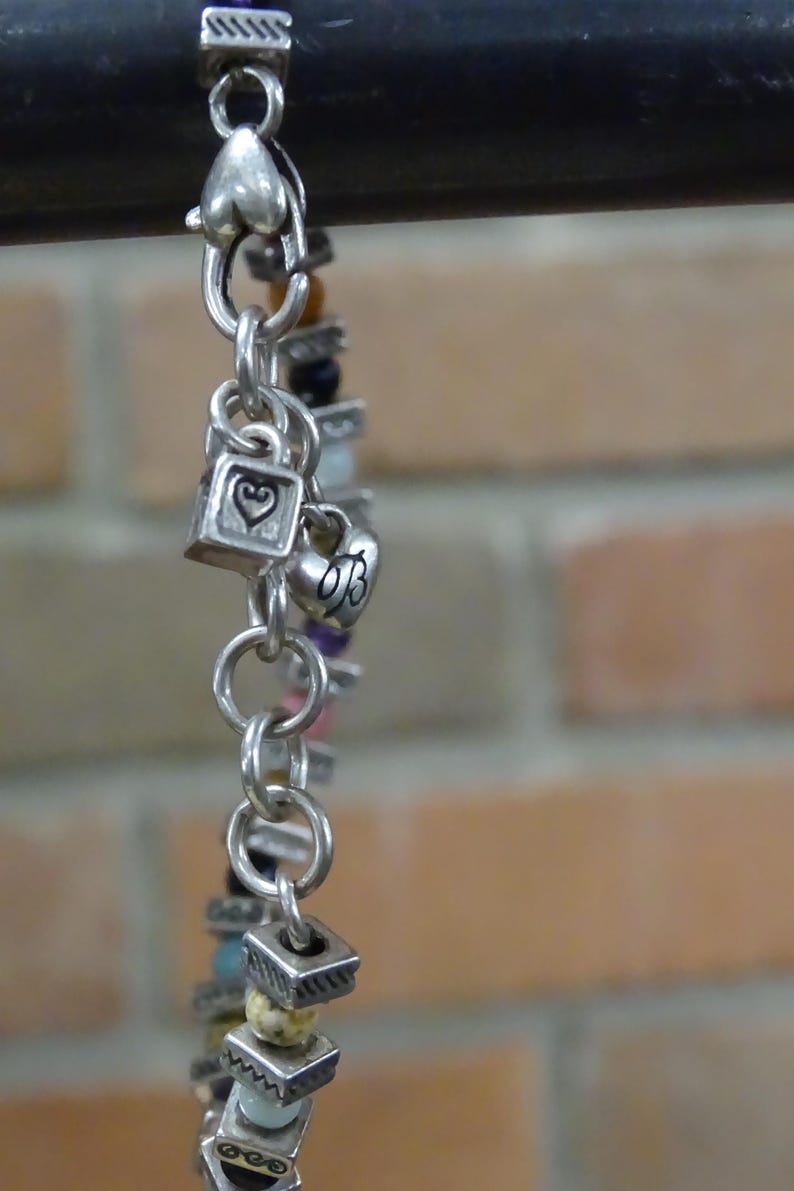 May include: Silver-toned charm bracelet with heart and square charms, and colorful beads. The bracelet features a variety of charms, including hearts and squares, and is strung with a mix of colorful beads. The bracelet is displayed against a brick background.