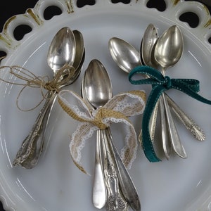 Op de afbeelding: Een set van zes zilveren lepels die met touw, jute en fluwelen lint aan elkaar zijn gebonden. De lepels zijn gerangschikt op een wit bord met een gegolfde rand.