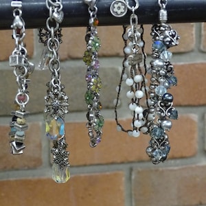 May include: A collection of six beaded bracelets with various designs and colors. The bracelets are silver-toned and feature charms, beads, and crystals. They hang from a black metal bar against a brick wall.