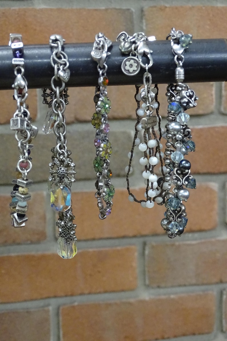 May include: A collection of six beaded bracelets with various designs and colors. The bracelets are silver-toned and feature charms, beads, and crystals. They hang from a black metal bar against a brick wall.