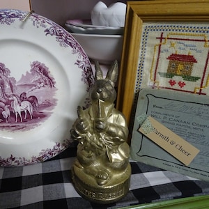 Puede incluir: Una figurita de conejo de color dorado que sostiene una pipa, sentada sobre una superficie a cuadros blancos y negros. La figurita está etiquetada como "Peter Rabbit".