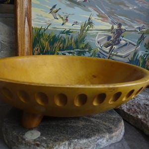 May include: A handcrafted, golden-yellow wooden bowl with a shallow, rounded design. The bowl features a series of evenly spaced, oval-shaped cutouts around the rim and rests on three small, rounded feet. The bowl is displayed on a gray stone.