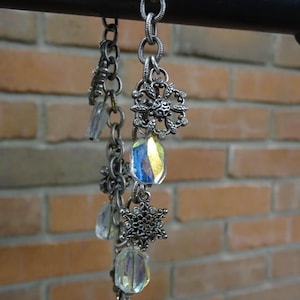 May include: A silver-toned chain bracelet with decorative snowflake and floral charms. The bracelet features iridescent, rectangular crystal beads. The background is a brick wall.