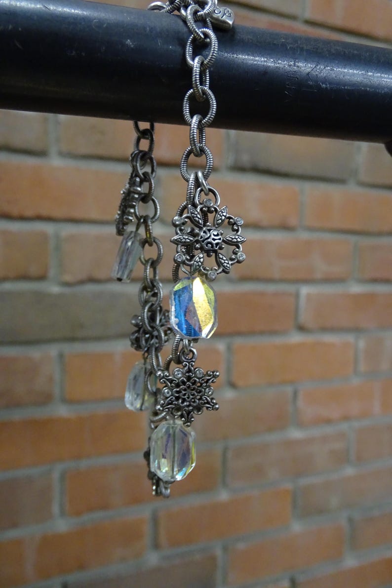 May include: A silver-toned chain bracelet with decorative snowflake and floral charms. The bracelet features iridescent, rectangular crystal beads. The background is a brick wall.