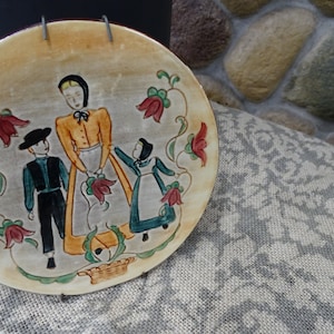 Puede incluir: Un plato de cerámica con un diseño pintado a mano de una familia de tres personas con ropa tradicional. La mujer lleva un vestido amarillo y un delantal blanco, el hombre lleva un traje negro y un sombrero negro, y la niña lleva un vestido azul y una capucha blanca. El plato está decorado con flores rojas y verdes.