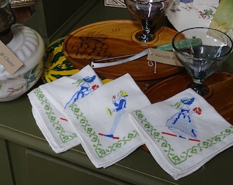 Op de afbeelding: Drie witte linnen servetten met geborduurde ontwerpen van mensen in blauwe en rode kleding. De servetten zijn gevouwen en gerangschikt op een houten oppervlak. De tekst "Warmth & Cheer" is zichtbaar op een label.