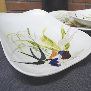 May include: White ceramic serving platter with a unique, wavy shape. The platter features a colorful botanical design with green, yellow, and blue accents. Additional matching plates are visible in the background. This vintage dishware is perfect for serving.