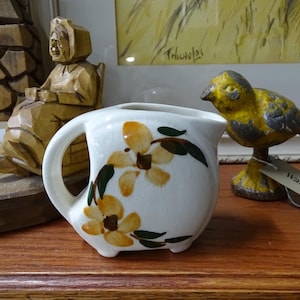 May include: A white ceramic creamer with a floral design featuring yellow flowers and green leaves. The creamer has a handle and a spout.
