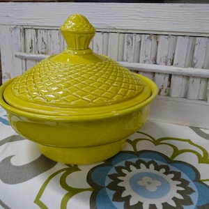 May include: A bright yellow ceramic serving dish with a lid. The lid has a textured diamond pattern and a decorative finial. The dish sits on a patterned tablecloth with blue, green, and gray floral designs. The background includes a white distressed shutter.